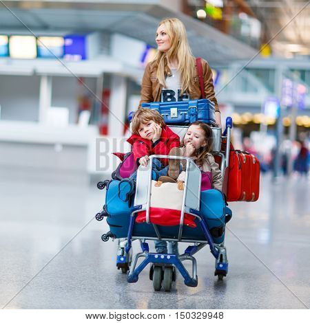 Two adorable little siblings, kid boy and girl sitting on suitcases on international airport. Young mother pushing. Brother and sister, happy family wating for flight and going on vacations.