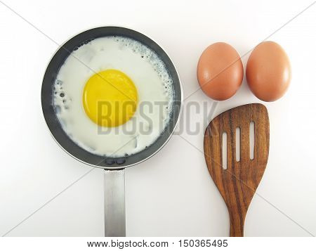 fried egg in small metallic teflon pan with silver stainless handle, two raw eggs and brown wooden spatula isolated on white background, preparation cooking breakfast, flat lay close-up top view