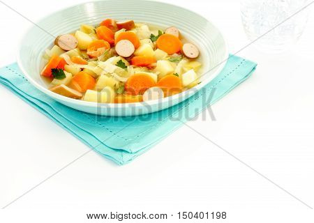 Vegetable Stew with sliced Frankfurter sausage in a blue soup bowl on top of aqua blue napkin and a glass of water with ice cubes on the right side. On white background with lots of copy space.
