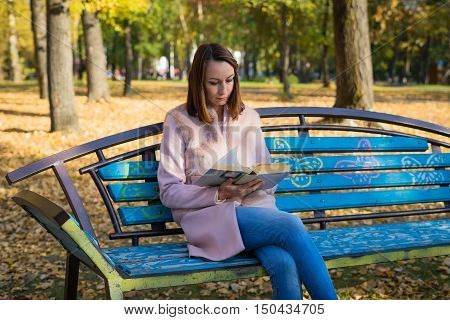 Girl sits on a park bench and reading a book sunny autumn day in October. Yellow leaves underfoot and the trees.