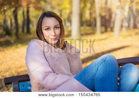 Girl sits on a park bench sunny autumn day in October. Yellow leaves underfoot and the trees.