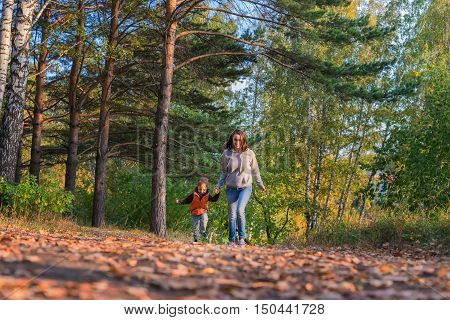 Mother and son running on path in autumn forest. Family at nature autumn bright sunny day fallen yellow leaves underfoot.