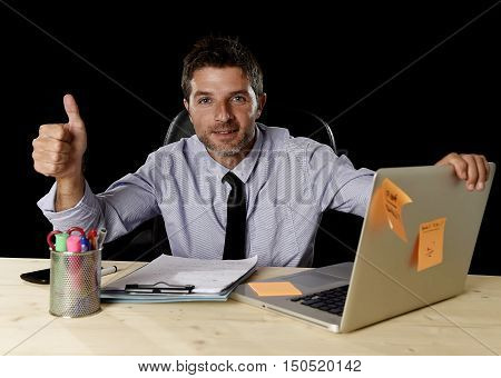 corporate portrait of happy successful businessman in shirt and tie smiling at office desk working with laptop computer isolated on black background confident and trustful in business success concept