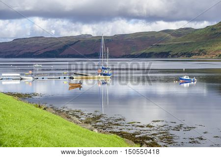Boats and their reflections on the beautiful calm waters of Loch Harport at Carbost Isle of Skye Scotland with hills of purple heather beyond under a cloudy sky