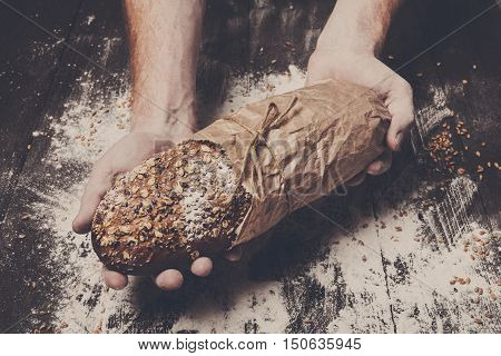 Warm fresh rye whole grain bread, wrapped in craft paper. Healthy baking and cooking concept background. Hands carefully hold loaf on wooden table background, sprinkled with flour. Soft toning