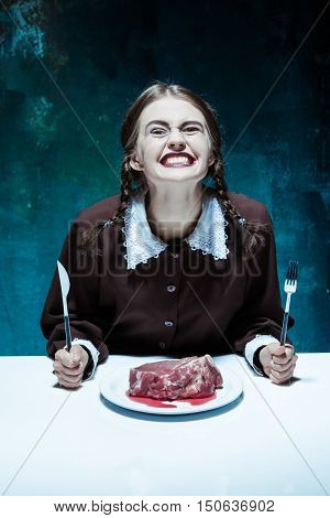 Portrait of a young girl with knife, fork and and a piece of raw meat on a plate. Girl in school uniform as killer. The image in the style of Halloween and Addams family