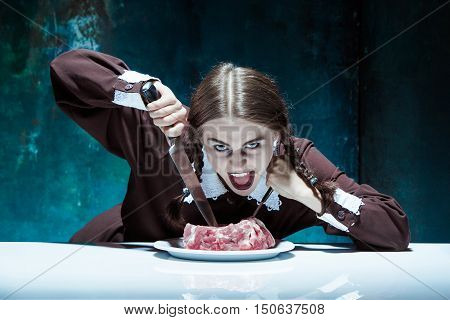 Portrait of a young girl with knife fork and and a piece of raw meat on a plate. Girl in school uniform as killer. The image in the style of Halloween and Addams family