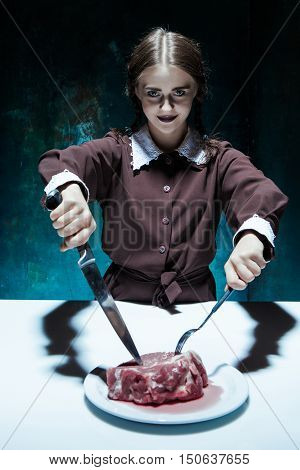 Portrait of a young girl with knife fork and and a piece of raw meat on a plate. Girl in school uniform as killer. The image in the style of Halloween and Addams family