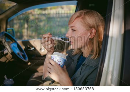 Girl Eating Ice Cream With A Spoon In The Car. Blond Girl In A Raincoat, Eats Ice Cream While In The