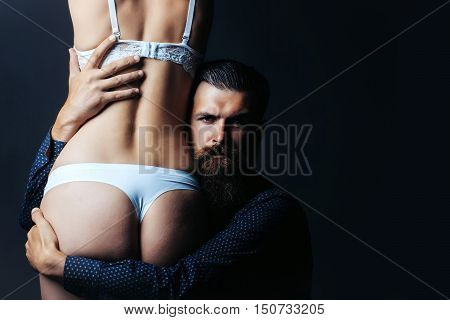 handsome bearded man with long beard on serious face embracing young woman with sexy body legs and buttocks with bare back in lingerie on grey background copy space