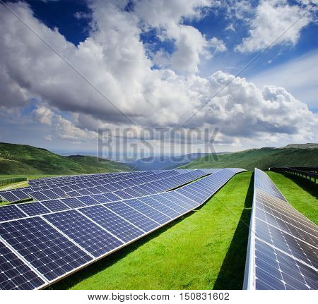 Beautiful modern solar station with blue panels standing in field with green grass and mountains at the horizon under a bright cloudy sky with reflection of clouds in batteries