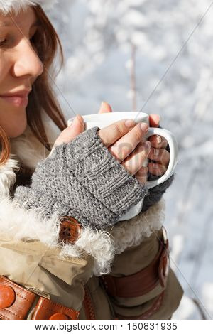 Beautiful young smiling woman in gray fingerless knitted mittens cap and coat with white fur holding in hands white cup of tea or coffee at the background of snowy forest. Closeup.