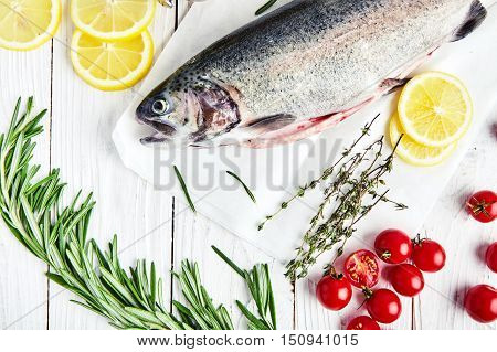 Ingrediets for baking trout: raw fresh rainbow trout on paper with cherry tomatoes, slices of lemon, sprigs of rosemary, thyme, pepper mill. Preparations for cooking delicious fish. White wood back