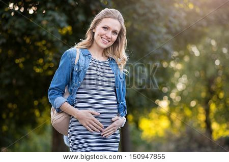 Waiting for a baby.Pretty pregnant woman smiling and stroking belly while walking in the park.
