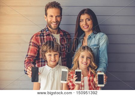 Beautiful young parents and their children are holding smartphones looking at camera and smiling on gray wall background