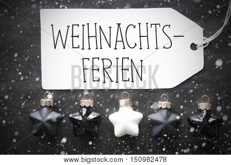 Label With German Text Weihnachtsferien Means Christmas Holidays. Black And White Christmas Tree Balls On Black Paper Background With Snowflakes. Christmas Decoration Or Texture. Flat Lay View