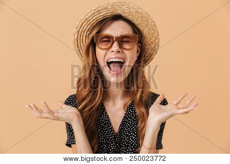 Photo of unhappy capricious woman 20s wearing straw hat and sunglasses throwing arms aside and screaming isolated over beige background