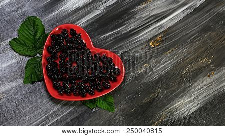 Organic Black Raspberry With Leaves On A Decorative Red Plate In The Form Of A Heart, On A Dark Back