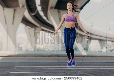 Horizontal Shot Of Sportive Beautiful Young Female In Purple Top And Sneakers, Holds Jump Rope, Does