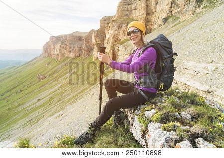 Smiling Girl Traveler In A Yellow Hat And A Pair Of Sunglasses Ssitting At The Foot Of Epic Rocks Wi