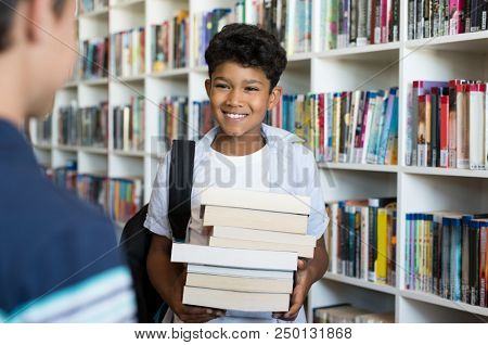 Young arab boy holding stack of books and talking to classmate in library. Happy two children have a conversation. Middle eastern child discussing study with his friend in a primary school library. 