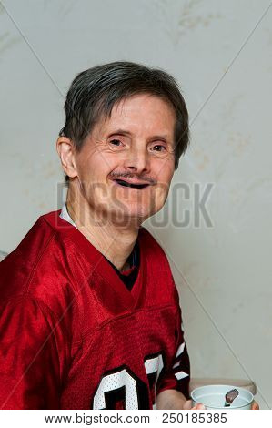 Portrait Of An Elderly Man With Downs Syndrome.  He Is Holding A Cup With A Spoon In It And Smiling 