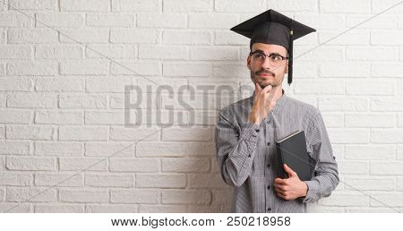 Young adult man over brick wall wearing graduation cap serious face thinking about question, very confused idea