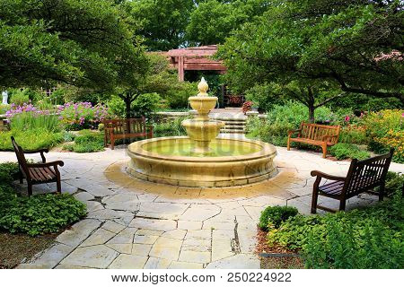 Courtyard Including A Water Fountain Surrounding Chairs Besides Manicured Plants And Trees Taken In 