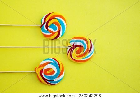 Colorful Hard Candy Lollipop On Yellow Pastel Background. Rainbow Sweet Pattern On Stick Flying Arou