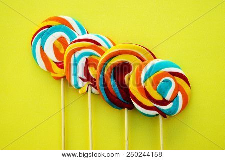 Colorful Hard Candy Lollipop On Yellow Pastel Background. Rainbow Sweet Pattern On Stick Flying Arou