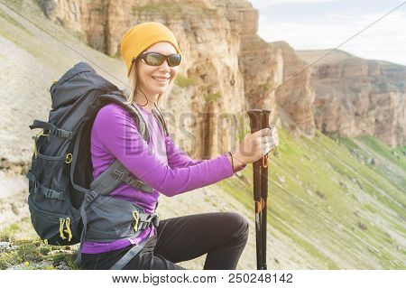Smiling Girl Traveler In A Yellow Hat And A Pair Of Sunglasses Ssitting At The Foot Of Epic Rocks Wi
