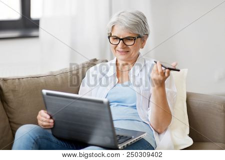technology, old age and people concept - happy senior woman in glasses with laptop computer and smartphone using voice command recorder at home