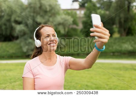 fitness, technology and healthy lifestyle concept - smiling woman in headphones listening to music and taking selfie by smartphone at park