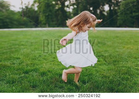 Pretty Little Cute Child Baby Girl In Light Dress Circling Around And Have Fun On Green Grass Lawn I