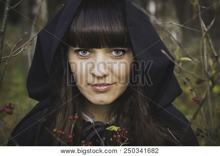 The Girl Is A Witch Is A Witch In A White Robe, And Black Coat In The Autumn Forest Reading A Spell 