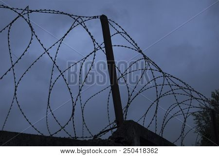 Barbed Wire At The Top Of The Fence Against The Background Of A Dramatic Sky. Without Color