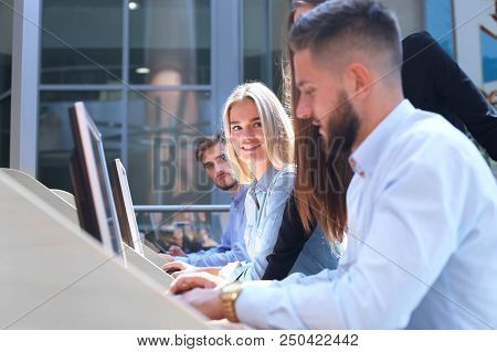 Group Of Young People In Casual Wear Sitting At The Office Desk And Discussing Something While Looki