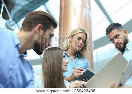 Group Of Young People In Casual Wear Sitting At The Office Desk And Discussing Something While Looki