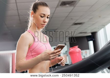 Slim Beautiful Girl With A Ponytale Standing On The Treadmill In The Gym Holding A Phone Wearing Ear
