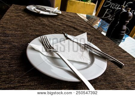 White Porcelain Plate, Knife, Fork, Paper Napkin, Saltpeter Set On The Street Restaurant Table With 
