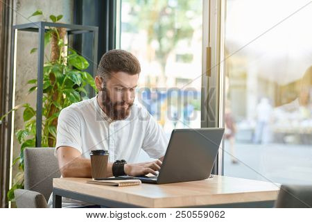 Young Man Working With Laptop Sitting Near Panoramic Windows. Drinking Coffee Gron Paper Can, Wearin
