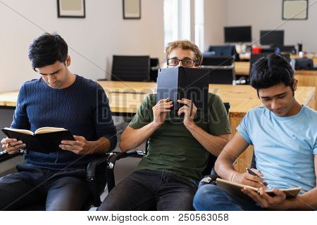 Student Hiding Behind Notebook And Sitting Between Two Friends. Three Fellow Students Preparing For 