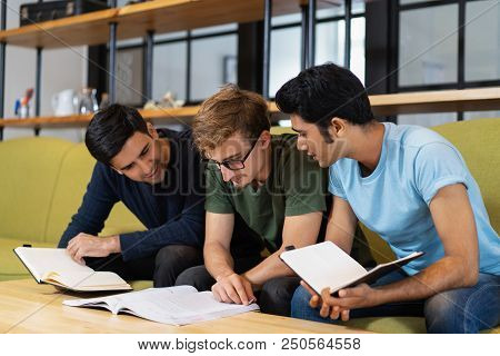 Three Fellow Students Reading Textbook And Preparing For Exam Together. Young Men Sitting On Couch A
