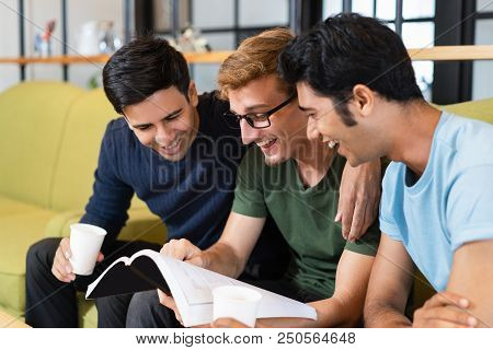 Three Fellow Students Reading Textbook, Laughing And Drinking Coffee Together. Young Men Sitting On 