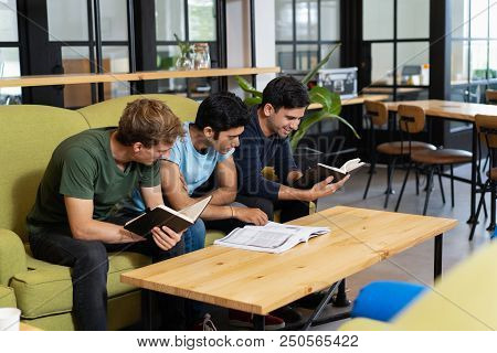 Three Fellow Students Studying And Preparing For Exam. Young Men Sitting On Couch In Library Reading
