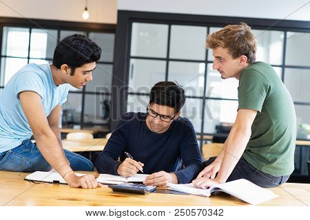 Three Serious Fellow Students Doing Homework Together. Young Men Sitting On Desk And Discussing Task