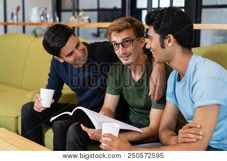 Three Smiling Fellow Students Reading Textbook And Drinking Coffee Together. Young Men Chatting And 