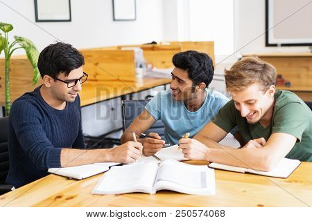 Three Students Studying And Doing Homework Together. Young Men Talking, Writing And Sitting At Desk 