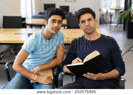 Two Serious Fellow Students Studying And Looking At Camera. Young Men Sitting In Armchairs In Classr
