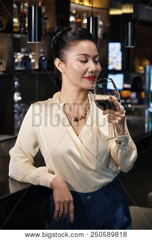 Gorgeous Young Asian Delighted Female Standing Relaxed Leaning On Dark Bar Counter And Smelling Red 